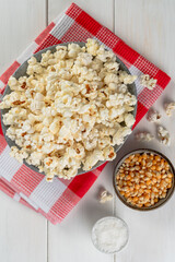 bowl with popcorn, popcorn kernels and salt on a red checkered napkin, on a white wooden table, top view.