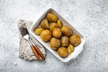 Hasselback potatoes with herbs in white ceramic casserole dish on white concrete table background...