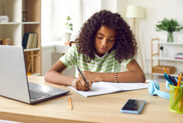 Diligent school child studying at home. Pretty African American girl sitting at desk, doing homework, writing essay in her notebook, using modern laptop and learning a lot of new information - obrazy, fototapety, plakaty