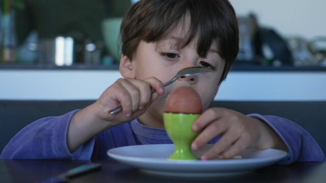 One small boy trying to open top of egg at breakfast table. Kid eating oeuf a la coque. Funny child nutritious protein food