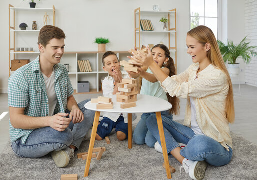 Friendly happy family with two children are playing tumbling tower sitting on floor at home in weekend. Mom, dad, son and daughter are spending time together. Family lifestyle, love and care concept.