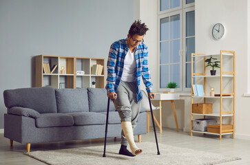 Young African American man with a broken leg in a cast walking with crutches in the living room at...