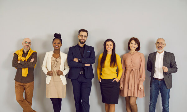 Portrait of multiracial representatives of modern business team standing in row near wall. Smiling men and women of all ages and nationalities dressed in smart casual clothes posing near gray wall.
