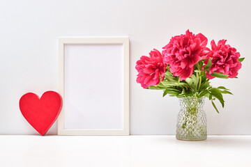 Mockup with a white frame and red peonies in a vase, red heart on a white table. Empty poster frame mockup for presentation design, text, lettering. Valentines Day, Happy Women's Day concept