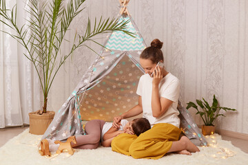 Image of beautiful brown haired woman sitting on floor near children's teepee and talking cell phone while her daughter sleeping on her legs, female having conversation. © sementsova321