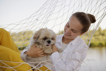 Outdoor shot of smiling happy joyful Caucasian woman wearing white shirt lies in a hammock with Pekingese dog on the bank of the river, female resting with her pet near the lake. - Powered by Adobe