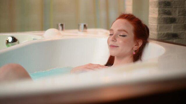 Close up of happy redhead female sitting in whirlpool with closed eyes and enjoying hydromassage at luxury spa salon. Beautiful young woman enjoying procedure of hydromassage. Shooting in slow motion.