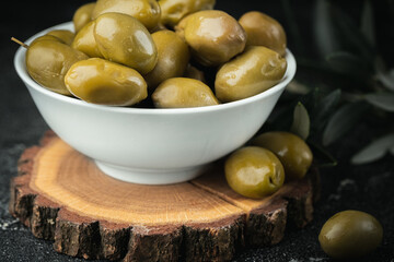 Close up shot of green olives in a white bowl on the wooden stand with olive leaves on a black background. Traditional Greek and Italian food.