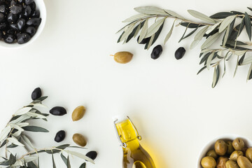 Green and black olives in white bowls next to a bottle with olive oil and leaves on a white...