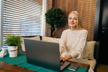 Portrait of charming young woman with long blonde hair sitting at table in cafe while using laptop computer and smiling looking at camera. Friendly freelancer using online application working remote