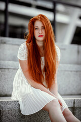 a red-haired girl in a light skirt; a very cheerful girl with long red hair; lips painted with red lipstick; sits on a bench and has a good mood; poses for a photographer; street photo session 