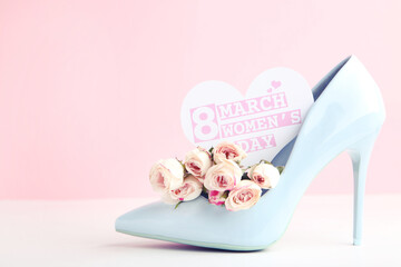 Flowers of roses and card in shape of heart with text 8 March Women's Day and blue high-heeled shoe on pink background