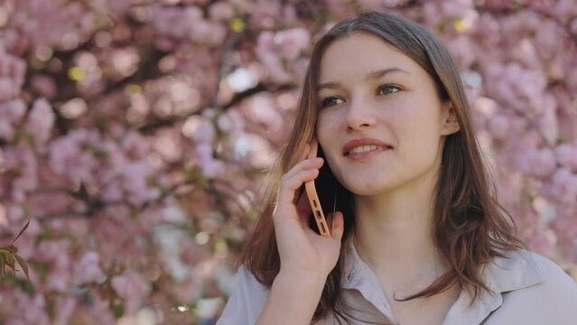 Charming young caucasian woman enjoying pleasant mobile conversation talking smartphone while standing outside near sakura tree with pink blooming. Concept of people and technology communication
