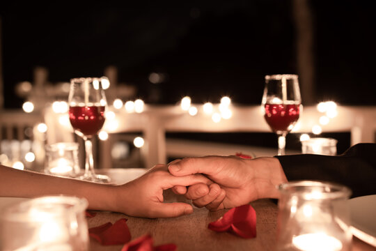 Romantic candlelight dinner table setup for couple with beautiful light as background. Man & Woman hold hands and drink wine. Concept for valentine's day and date.
