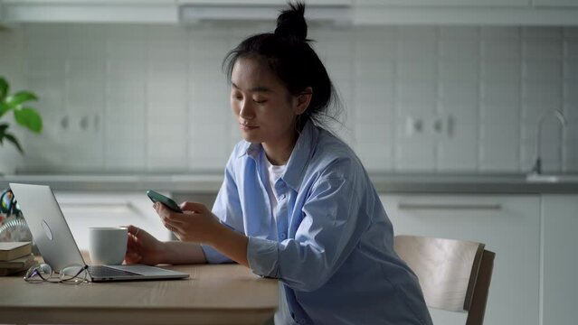 Procrastinating uninitiative Asian woman browsing social media feed on phone sits at kitchen table with laptop. Unmotivated Korean girl freelancer is unwilling to do work of digital addiction