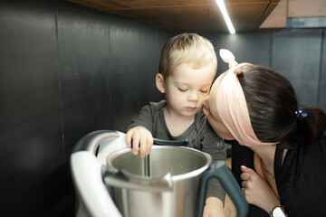 Mom with love kiss little son on cheek. Baby look at deep metal bowl of blender. Little child help...
