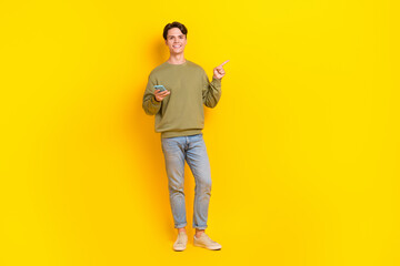 Full length photo of cheerful guy hold smart phone indicate finger empty space isolated on yellow color background