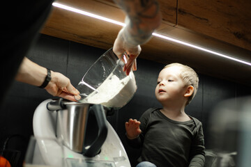 Pour flour from deep glass bowl into blender. Little boy help dad cook dinner. Baby in kitchen among modern appliances.