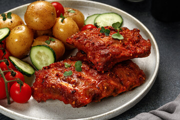 Plate with Marinated pork bbq ribs, young potatoes, tomatoes on black dark background. Hearty Dinner, lunch, meal.