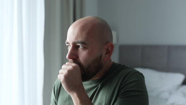 Sad bald unshaven man coughing, covering mouth with hand, touching throat, sitting on bed and looking out light window