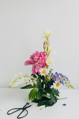 Stylish peony, lupin, iris and daisy arrangement on kenzan with scissors on rustic wood. Creative floral image. Modern summer flowers composition on rustic white table indoors.