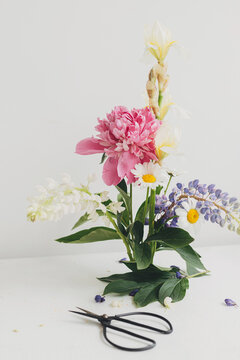 Stylish peony, lupin, iris and daisy arrangement on kenzan with scissors on rustic wood. Creative floral image. Modern summer flowers composition on rustic white table indoors.