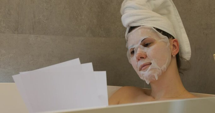 Woman with moisturizing sheet mask for face lie in bath and read. Beautiful female wearing towel on head in bathroom.