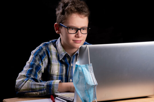Boy with curls and glasses and checked shirt works intently on laptop. Mouth guard, mask and fountain pen are also ready.