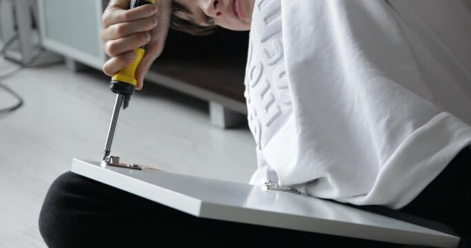 Unrecognizable teenage boy tightening screw in hinge of door of cabinet with yellow screwdriver, assembling furniture.