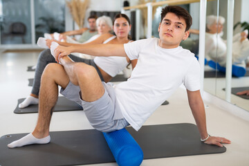 Sporty slender males and females doing exercises with pilates roller during group training at gym. Healthy lifestyle concept.
