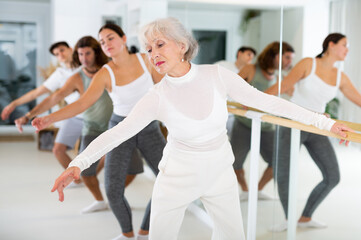 Gracile old-aged woman engaging in ballet at ballet barre in training hall during workout session