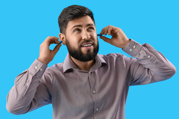 Young man with ear plugs suffering from loud noise on blue background