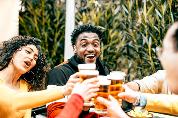 Group of friends having fun and drinking beer at bar restaurant - Multi ethnic people sitting at pub table toasting lager glasses in brewery garden - Happy hour party lifestyle concept
