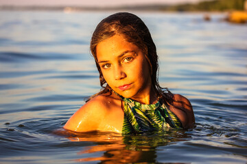 A beautiful young serious teenage girl posing in a sea. Portrait of a pretty female kid, schoolgirl in a calm water on sunset looking at a camera. Summer holiday at a children's camp. Fun, joy, relax.