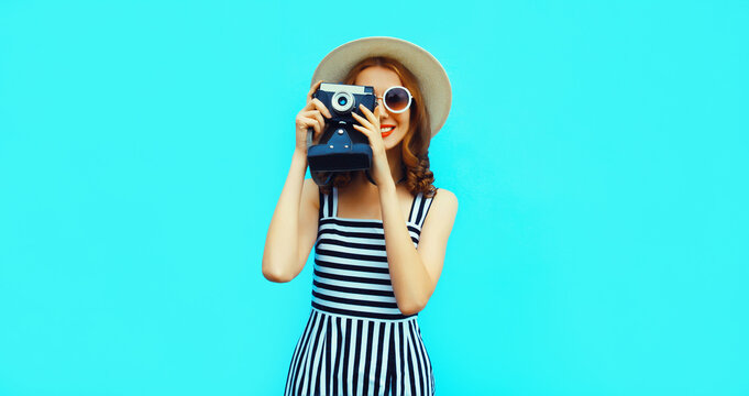 Summer portrait of happy smiling young woman photographer with vintage film camera wearing straw hat, dress, sunglasses on blue background