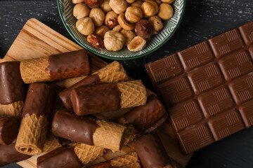 Board of delicious wafer rolls, chocolate bar and bowl of hazelnuts on grunge background