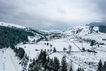 Aerial view of pine forest and a small village covered by snow in Carpathian Mountains of Romania, Rarau Mountains region. Cloudy weather conditions