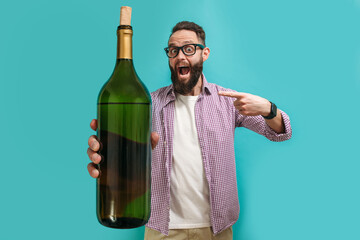 Young bearded charismatic hipster somilier showing a big bottle of wine, isolated on a blue studio...