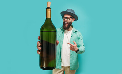 Young bearded charismatic hipster somilier showing a big bottle of wine, isolated on a blue studio...