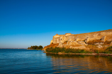Fototapeta premium Old stone fortress on the shore of the lake. Beautiful sunny landscape with fortress and blue sky