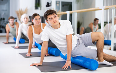 Diligent young man doing pilates exercises with roller on gray mat during workout session