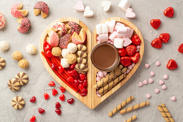 Valentine's Day concept. Top view photo of wooden heart shaped serving tray with confectionery scattered candies cookies and glass cup of hot chocolate on concrete texture background