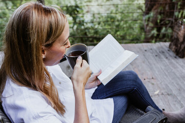 Hispanic woman reading a book in a  morning and holding coffee cup on the terrace of a country...