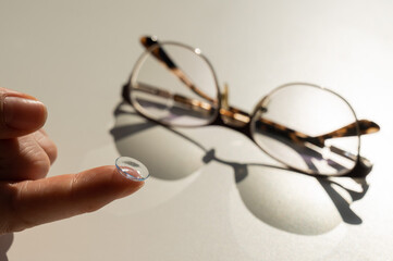 Close-up of a contact lens on a female index finger against the background of glasses on a white...