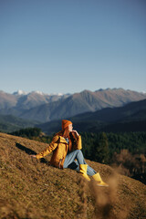 Woman full-length sitting resting on a hill and looking at the mountains in a yellow raincoat and jeans happy hiking trip in the autumn, freedom lifestyle in motion