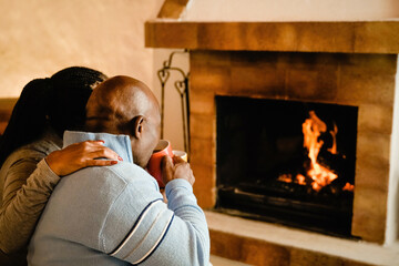 Senior African couple by cozy fireplace drinking hot chocolate together - Elderly people hugging...