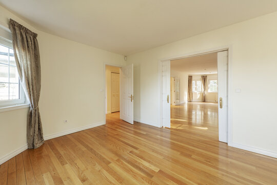 Dining room of a spacious empty house with wooden floors, sliding double doors, windows with curtains and access to multiple rooms