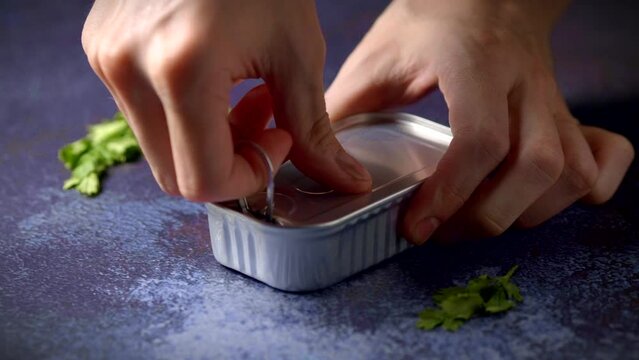 Hand opening a can of mackerel on a dark blue table with parsley. Ready to eat.