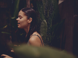 Fototapeta na wymiar A woman, a body in a swimsuit washes her head in a tropical shower outdoors against the backdrop of green tropical leaves, flowers and palm trees. Body and hair care, tanned skin, smile, vintage