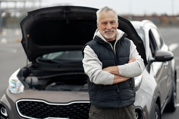 Confident senior man smiling at camera, standing with arms crossed against his car with open hood